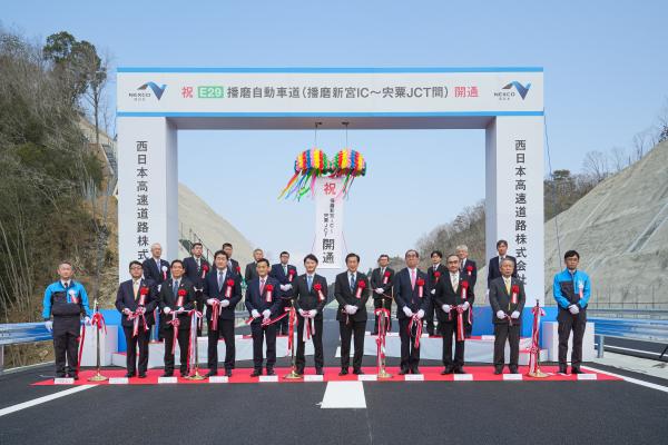 「祝 E29播磨自動車道（播磨新宮インターチェンジ～宍粟ジャンクション間）開通」と書かれた四角いアーチ門の前でスーツを着た沢山の関係者の方々がテープカットをしている写真