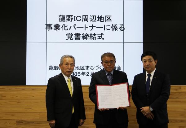 龍野インターチェンジ周辺地区事業化パートナーに係る覚書締結式と書かれたスクリーンの前で黒いスーツを着た3名の男性が並んでいる写真