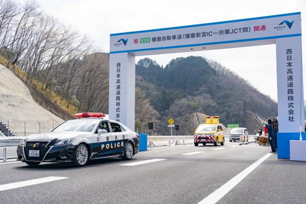 「祝 E29播磨自動車道（播磨新宮インターチェンジ～宍粟ジャンクション間）開通」と書かれた四角いアーチ門を警察車両を先頭に車が通っている写真