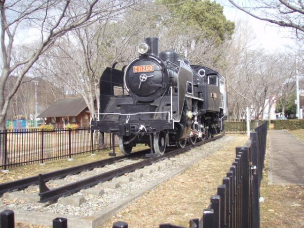 公園の中にある黒い柵の中に設置された黒色の蒸気機関車の写真