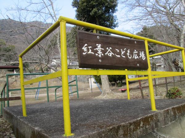 葉の落ちた木々の生えている公園を背景に、コンクリートブロックの上の黄色い鉄柵にかけられた木製の紅葉谷こども広場の看板の写真