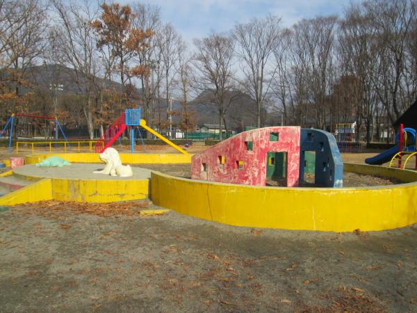 葉の落ちた木々を背景に、黄色の枠に囲まれた遊具のある砂場の写真
