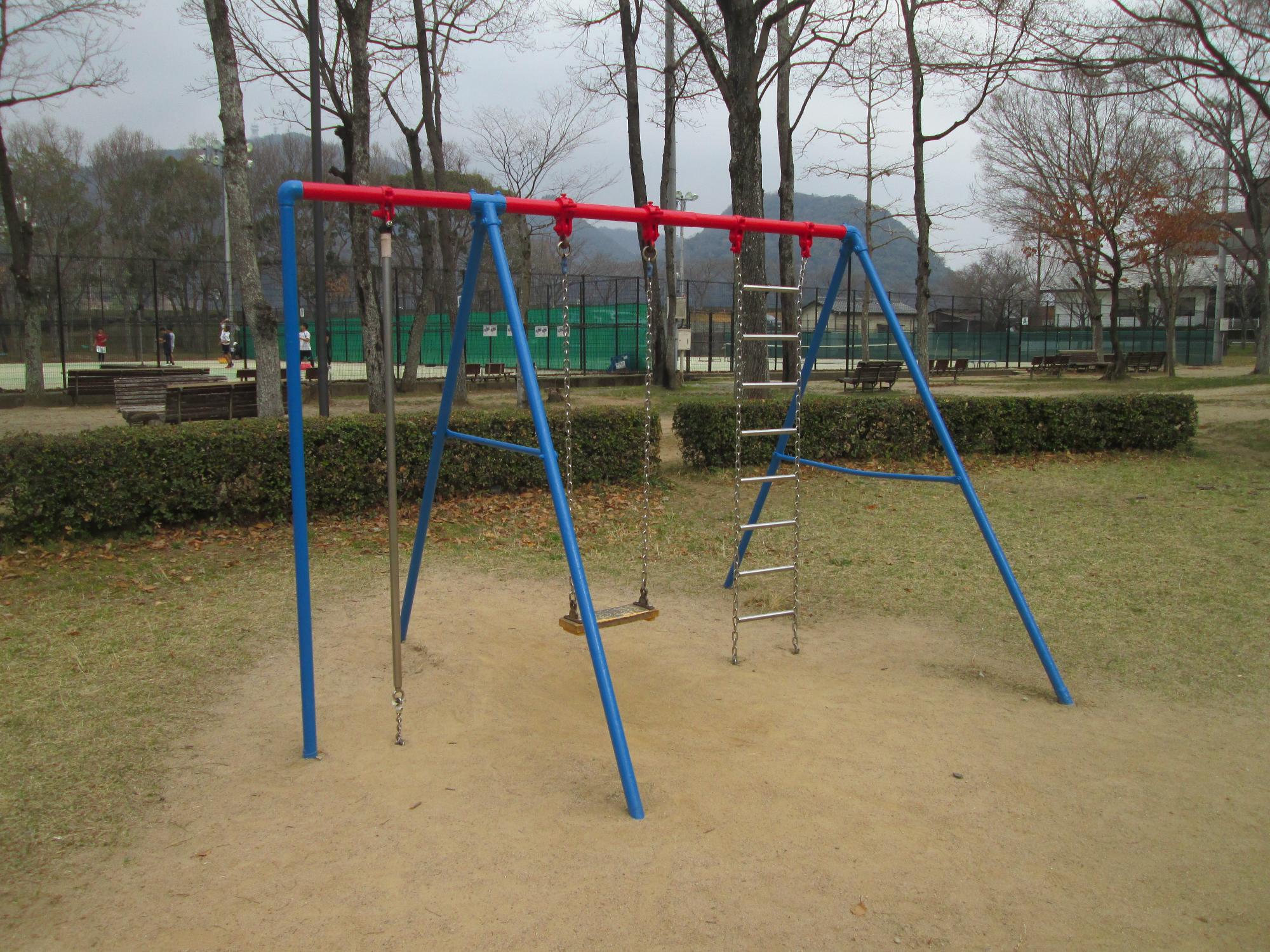 公園の中にある、トリコロールカラーのブランコ・チェーンネット・登り棒が一つになっている遊具の写真