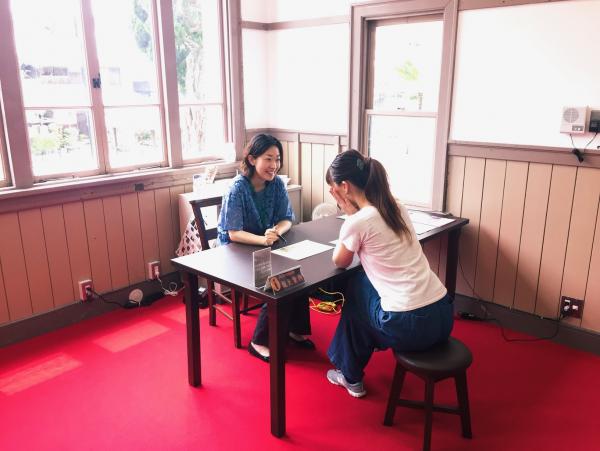 真っ赤な床に茶色のテーブルが置かれ向かい合わせに2人の女性が座って話をしている写真