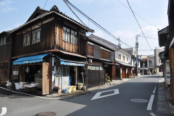 2階建ての瓦屋根の木造和風造りで、1階では青い屋根の下でお店を開けている家が建ち並ぶ今宿の町の写真