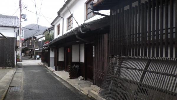 狭い路地の右側に白を基調とした2階建ての和風の古い町並みが続き、手前の家は窓から黒色の木の格子が設置された豆板ノ小路の写真