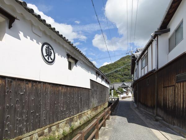 狭い通路の両側に下は黒の板張り上は白の外壁が続き、左側の外壁に黒で丸の中に東と書かれた看板が付けられた家が建つ小河町の写真