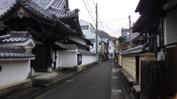 直線の狭い道路の左側には塀に瓦が載っている塀瓦の塀と、門に三角屋根に立派な瓦で造られている数寄屋門が設置された桶屋町の写真
