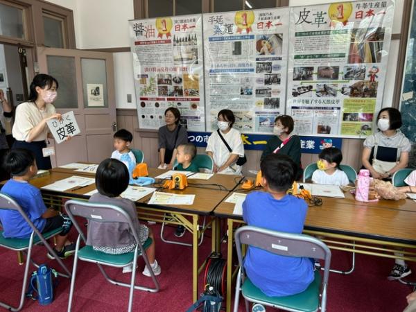 4ケの長机をくっつけパイプ椅子に子供たちが座り、真ん中の女性スタッフの説明を聞いている写真