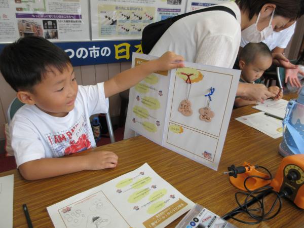 出来上がったキーホルダー2ケを用紙に貼り付け見せている男の子の写真