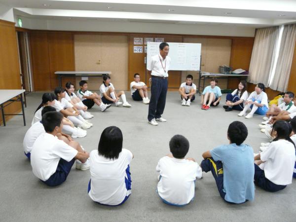 輪になって床に座る子どもたちと、その輪の中心に立ち子どもたちに向かって話をする男性の写真