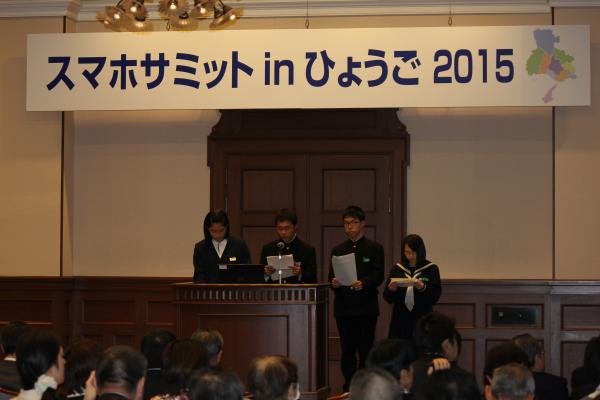 四人の学生さんたちが演台の前で立ち、前に座る大勢の人々の方を向いて発表をしている写真