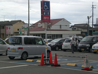 広い駐車場に車が止まっており、ピクトグラムの車いすマークや妊婦マークが記された看板が立っている写真