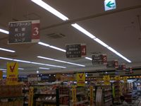 天井から種類ごとに分かれた吊り看板があり、食品別に品物が陳列された什器を通路側からみた写真