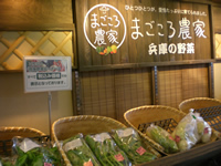 「まごころ農家」と書かれた看板が壁に掛けられており、竹かごの中に野菜が並べられている写真