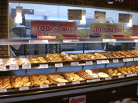 様々な種類のパンが2段に分かれて陳列されており、窓に「100円ベーカリー」の張り紙がされている写真
