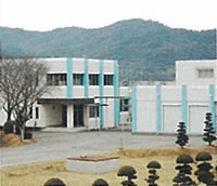 木々が植えられた広い敷地内に、水色の縦のラインが施された白壁の2棟の建物が設置された市場水源地の写真