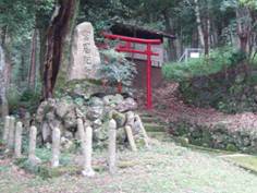 青々とした木々の生えている山の中で、石の土台の上にある石碑とその右後ろにある小さな赤い鳥居の写真
