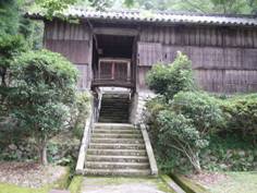 青々とした木々の生えている小さな石垣と階段の先にある、トタン屋根・木製の長屋の写真
