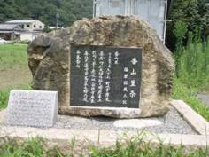 青々とした自然の場所に置かれている、石の土台の上に黒茶色が多めの石に埋め込まれた黒地に白で文字の書かれた石碑の写真