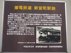 茶色の看板に黄色の文字で「播電鉄道 新宮町駅跡」とタイトルが書かれ、その下に白文字の説明と白黒の電車の写真がある写真