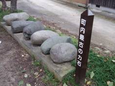 道の一角にある白色の文字で「町内最古の力石」と書かれた茶色の棒状の看板と、その脇にある長方形の石の台に並んでいる、6つの楕円形の石の写真