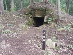 木々の生えている山の中にある縦長の木の棒の看板がある地下の石室のような場所に続く入り口の写真