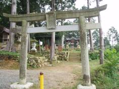 木々の生えている森のような場所にある石の鳥居の奥に祠のような物が見えている写真