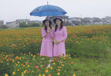 たくさんの紅花が咲いている場所に、青色の傘をさし、お揃いのピンクの衣装を着た女性2名が立っている写真