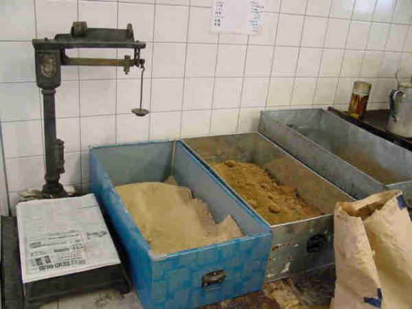 白いタイルの壁の作業場のような場所で、青や銀色の長方形の金属の入れ物に飴の材料になる粉のようなものが入れられて並べられてある写真