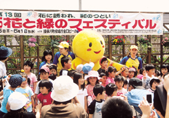 「花と緑のフェスティバル」と書かれた横断幕が貼られたイベント会場で、黄色のマスコットキャラクターの周りに沢山の子供たちが集まっている様子の写真