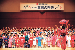 「第19回童謡の祭典」と書かれた横断幕が掲げられたステージ上で、浴衣を着た女性と向かい合って立つ大勢の子供たちが並んで立ち、両手を上げている音楽会の様子の写真