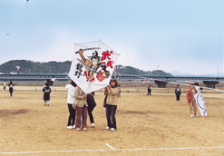 千鳥ヶ浜グランドの広場で参加者が凧あげを行っているなか、赤文字で「武蔵」黒文字で「龍野」と書かれ両手に二本の刀を持った武将のイラストを描いた六角形の大きな凧を、4人がかりで持っている童謡の里凧あげ祭りの様子の写真