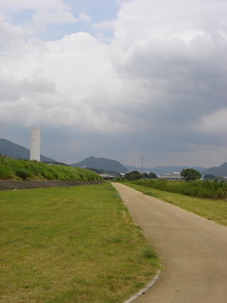 厚い雲がたちこめた空の下、左側に大きな白い煙突の様なものが立っている手前に、通路が真っすぐに整備された現在の揖保川の堤防の写真