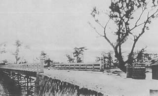 右側に一本の木が立つ木組みの樟津橋の上を人が渡っているモノクロ写真