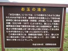 草原に立っている茶色の看板に黄色の文字で「お玉の清水」というタイトルとその下に白色で説明文が書かれている写真