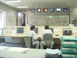 天井に6台のモニターが設置され、2人の作業員が座っている前にコンピューターが設置されている中央制御室内の写真
