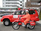 赤色のボディの車の傍に、ヘルメットを被りオレンジ色の救助服を着た消防士の男性が消防活動二輪車にまたがっている写真