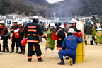 広場に並んだ白色のヘルメットを被った人たちが、消防士の指示のもと消火バケツを搬送しているバケツリレーの様子の写真