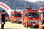 アーチの奥から進んでくる消防車に乗った消防士が、間隔をあけて立っている2名の制服姿の消防士を向いて敬礼を行っている出初式の様子の写真