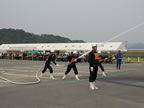 白色のテントが設置された広場の中央で、白いヘルメットを被りオレンジ色のゼッケンをつけた青色の作業服を着た消防士が真っすぐ前を向いて立ち、傍に立つ消防士が見守るなか2名の消防士が一緒にホースを持ち放水を行っている様子の写真