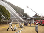 お寺の敷地内の広場に停車した消防車のはしごの上に乗った消防士や、防火衣を着た二人二組の消防士、防火服を着た消防士の方々がホースを持ち、隣接する山に向かって放水している消火訓練の様子の写真