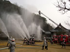 お寺の敷地内の広場に停車した消防車のはしごの上に乗った消防士や、防火衣を着た消防士、防火服を着た消防士の方々がホースを持ち、隣接する山や建物に向かって放水を行っている消火訓練の様子の写真