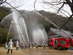 水浸しになったお寺の敷地内の広場で、消防車のはしごの上に乗った消防士や、防火衣を着た消防士、防火服を着た消防士の方々がホースを持ち、隣接する山に向かって放水を行っている様子を後方から撮影した写真