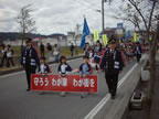 赤色の横断幕を持った3名の児童と制服姿の2名の消防士を先頭にパレードが行われている様子の写真