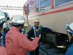 電車の車体が展示されている傍で、車体の下を指差している黄色のヘルメットを被った人と、白色のヘルメットを被りオレンジ色の救助服を着た消防士が話をしている訓練の様子の写真