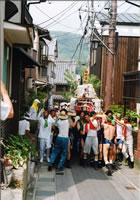 多くの人が担いでいる酒樽が乗ったみこしが路地を移動している写真