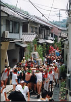 多くの人が担いでいる酒樽と魚の作り物を乗せた2基のみこしが路地を移動している写真