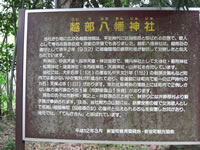 林のような場所にある茶色の看板にタイトルが黄色の文字で「越部八幡神社」と書かれその下に白い文字で説明が書かれている写真