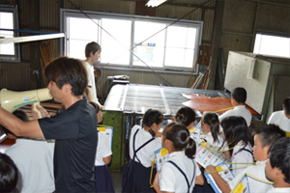 皮が広げられた机の横で、男性が拡声機を使って話している回りに資料を持った児童が集まっている写真